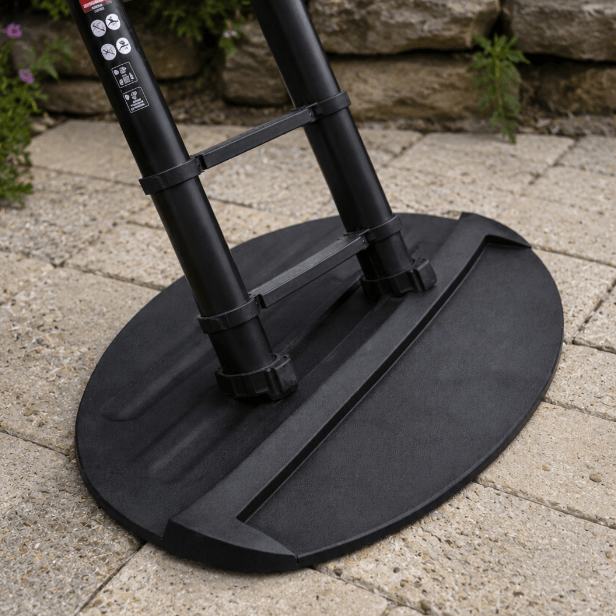 A close-up outdoor shot of a black round patio umbrella base and upper pole section resting on a light stone paver surface with a stone wall and small plants in the background.