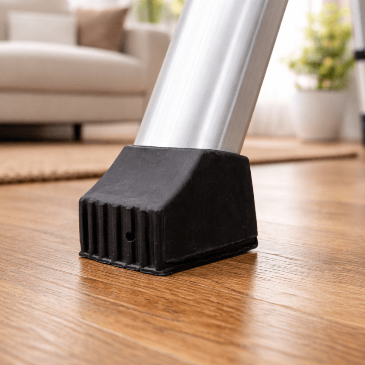 A close-up of a black plastic leg protector or floor guard with a ribbed triangular base, attached to a silver metal leg, resting on a wooden floor in a living room setting.