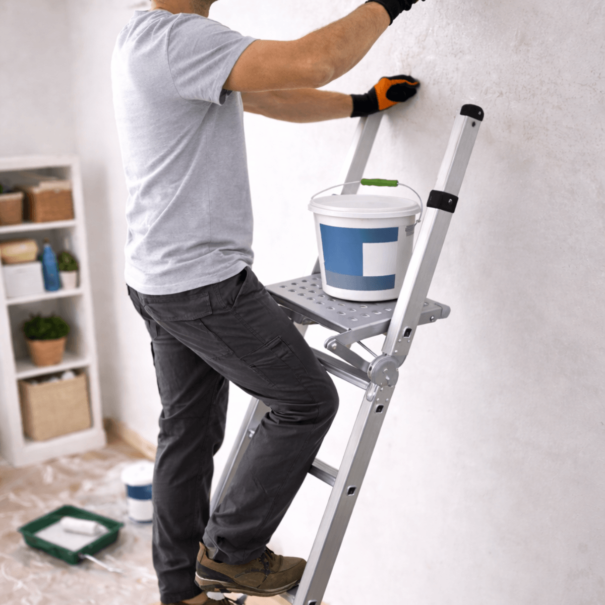 A man in a gray t-shirt and dark pants stands on a silver extension ladder, applying wallpaper or paint to a textured white wall in a room. A white paint bucket with a blue label sits on the ladder's platform.